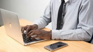A person in a gray dress shirt and tie types on a laptop at a desk with a smartphone resting beside it.