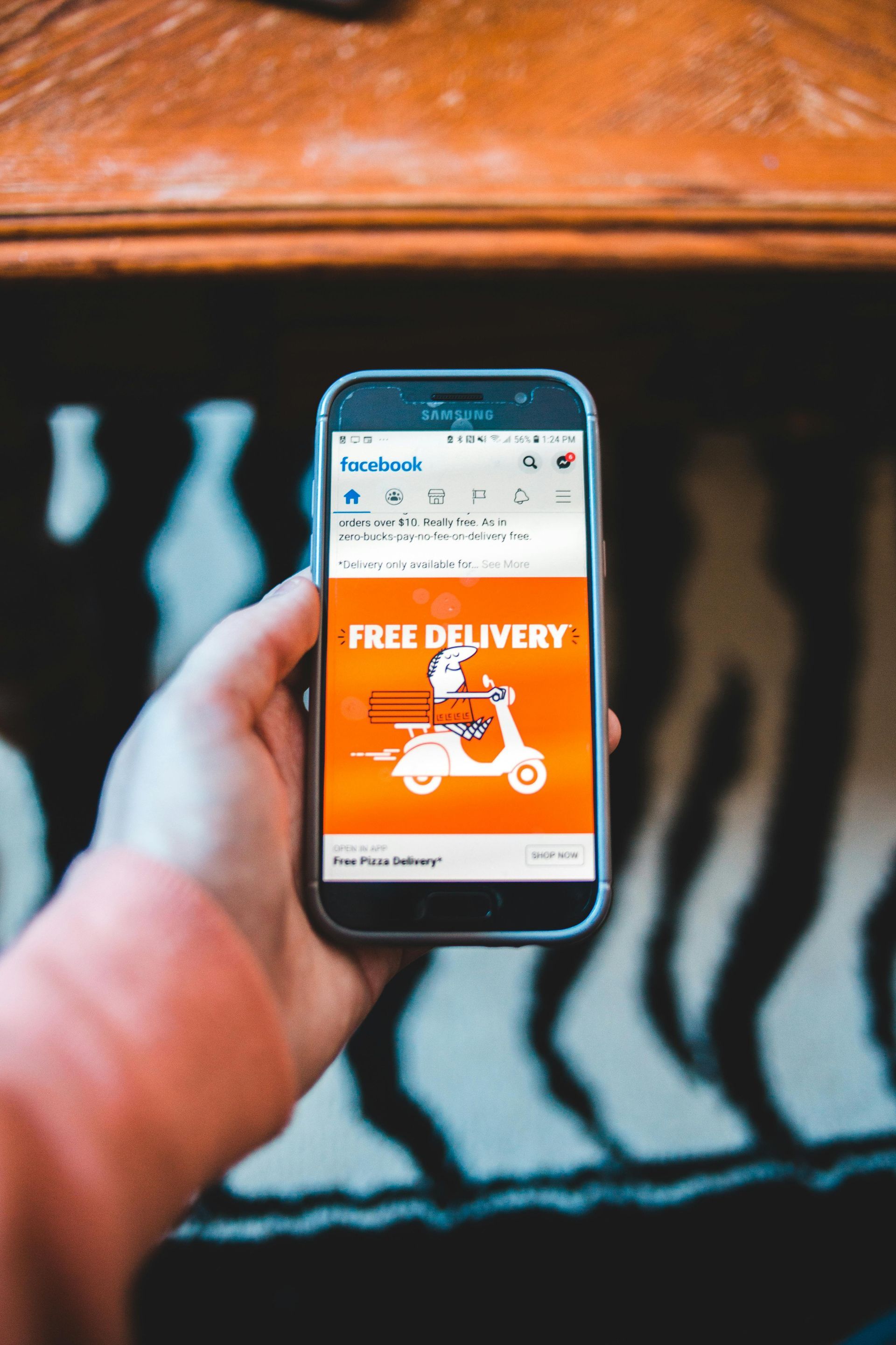 A hand holds a smartphone displaying a bright orange advertisement for free delivery featuring a scooter illustration.
