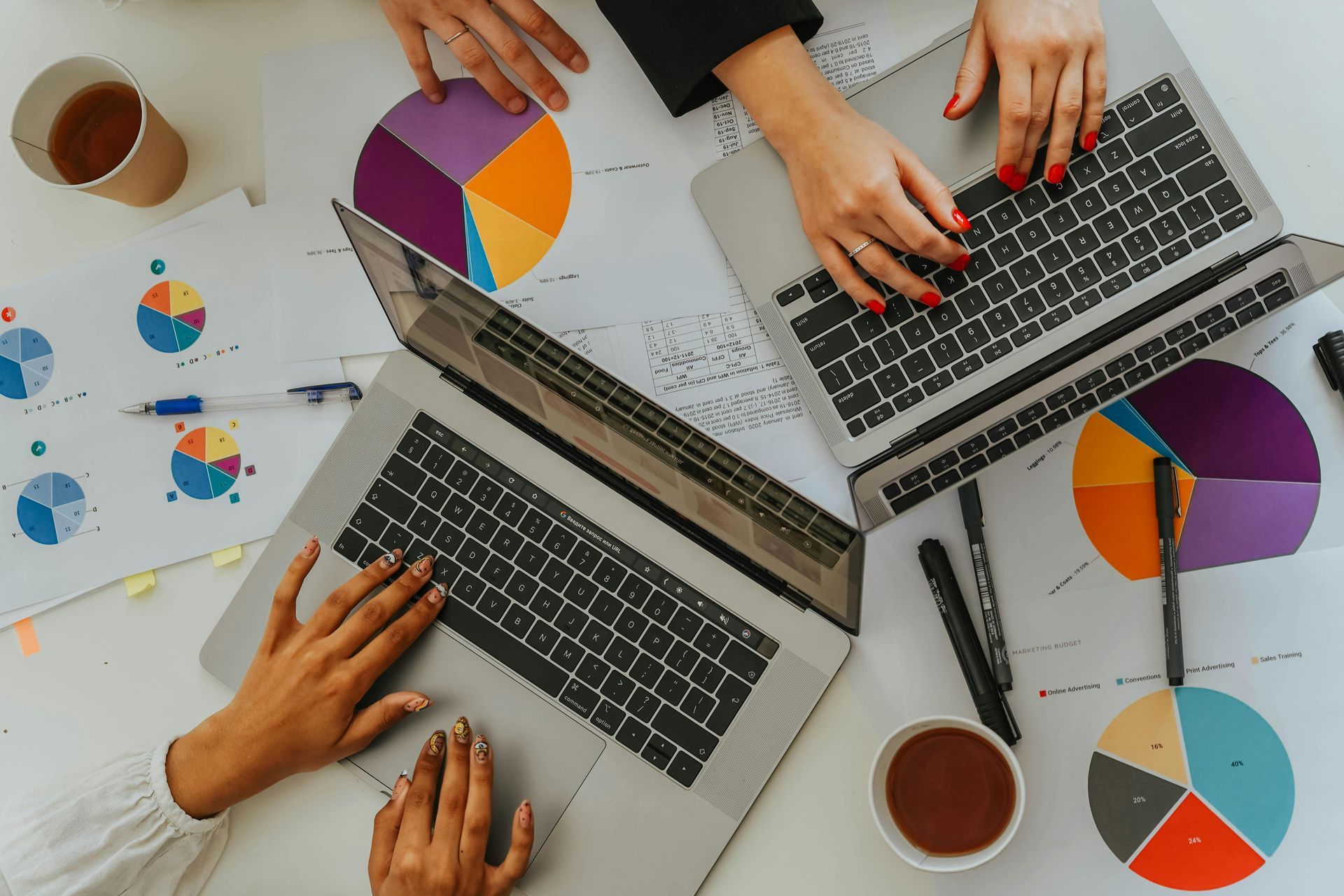 Two people use laptops on a desk covered with colorful pie charts, pens, and coffee cups.