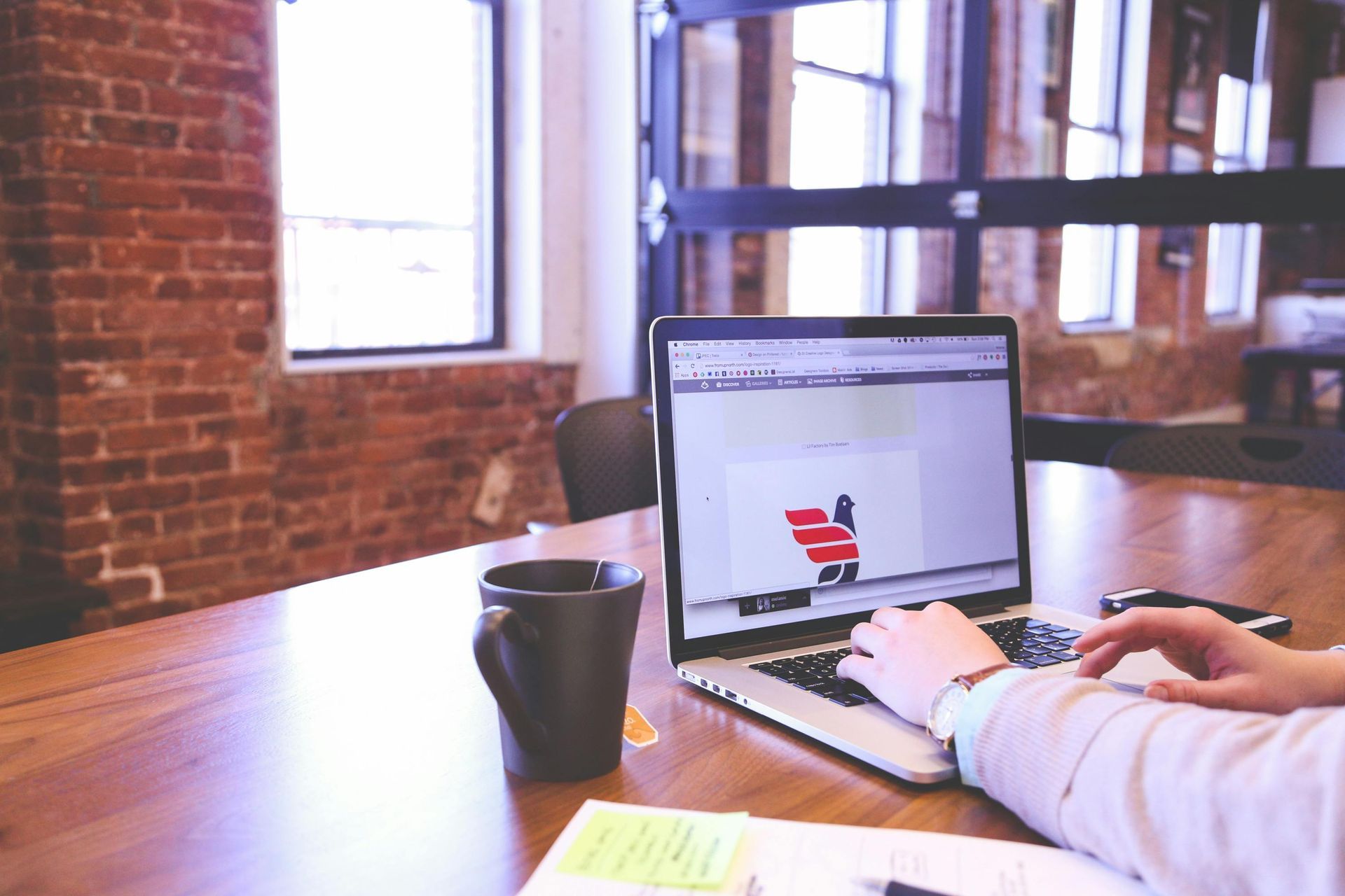 A person types on a laptop at a wooden table with a black mug, situated in an office with brick walls and large windows.