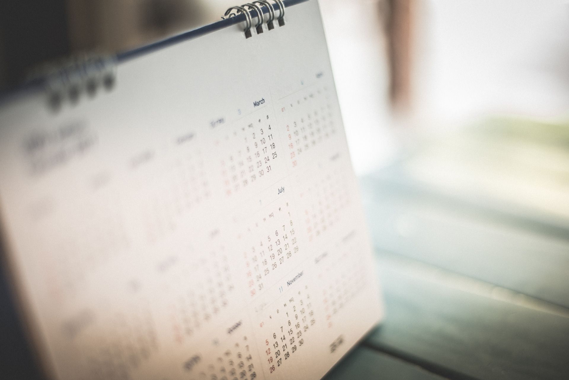 Close-up of a white desk calendar, resting on a wooden surface. The calendar shows multiple months.