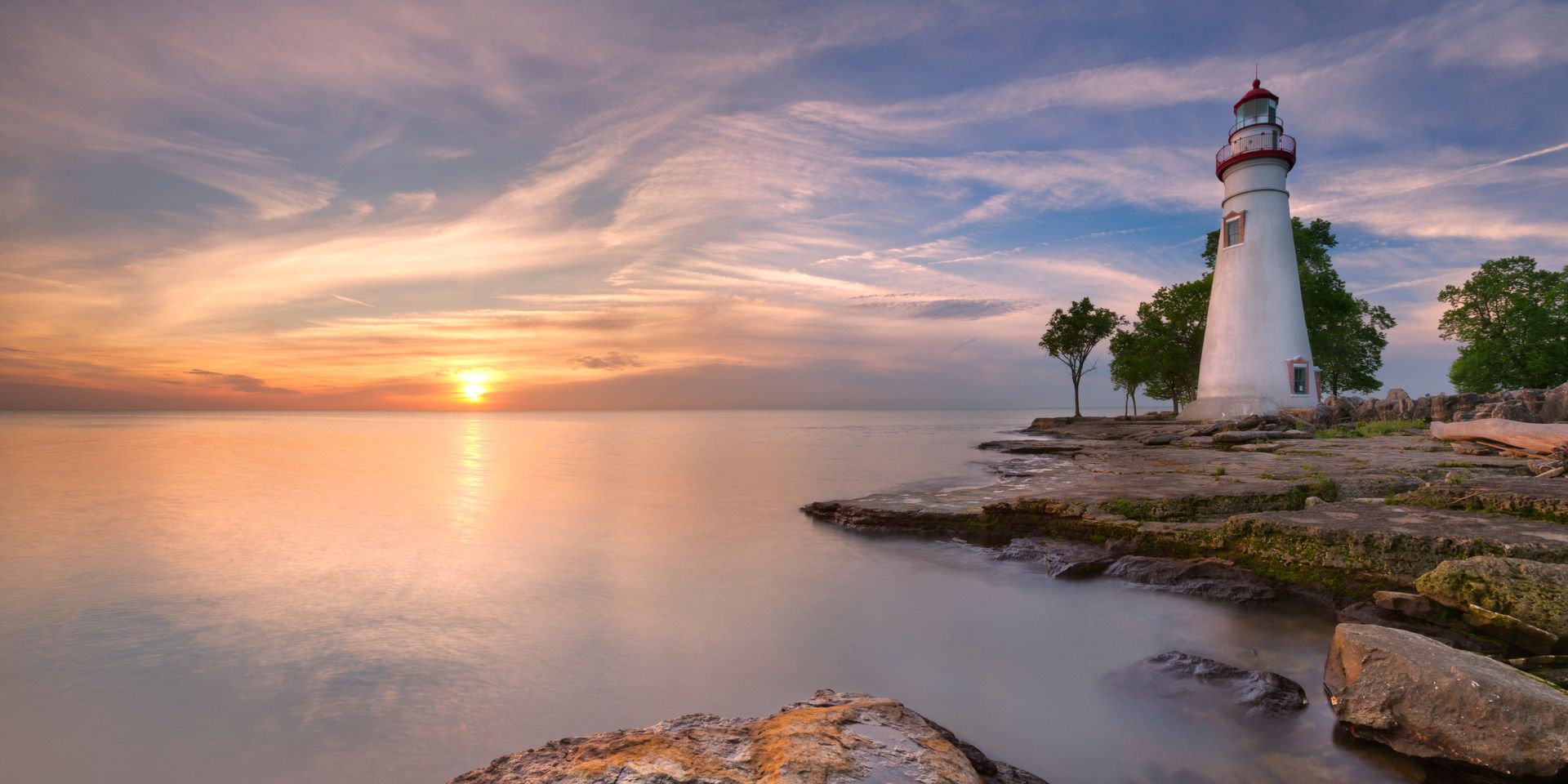 Lighthouse at sunset, white structure with red top on a rocky shoreline, orange and pink sky over calm water.