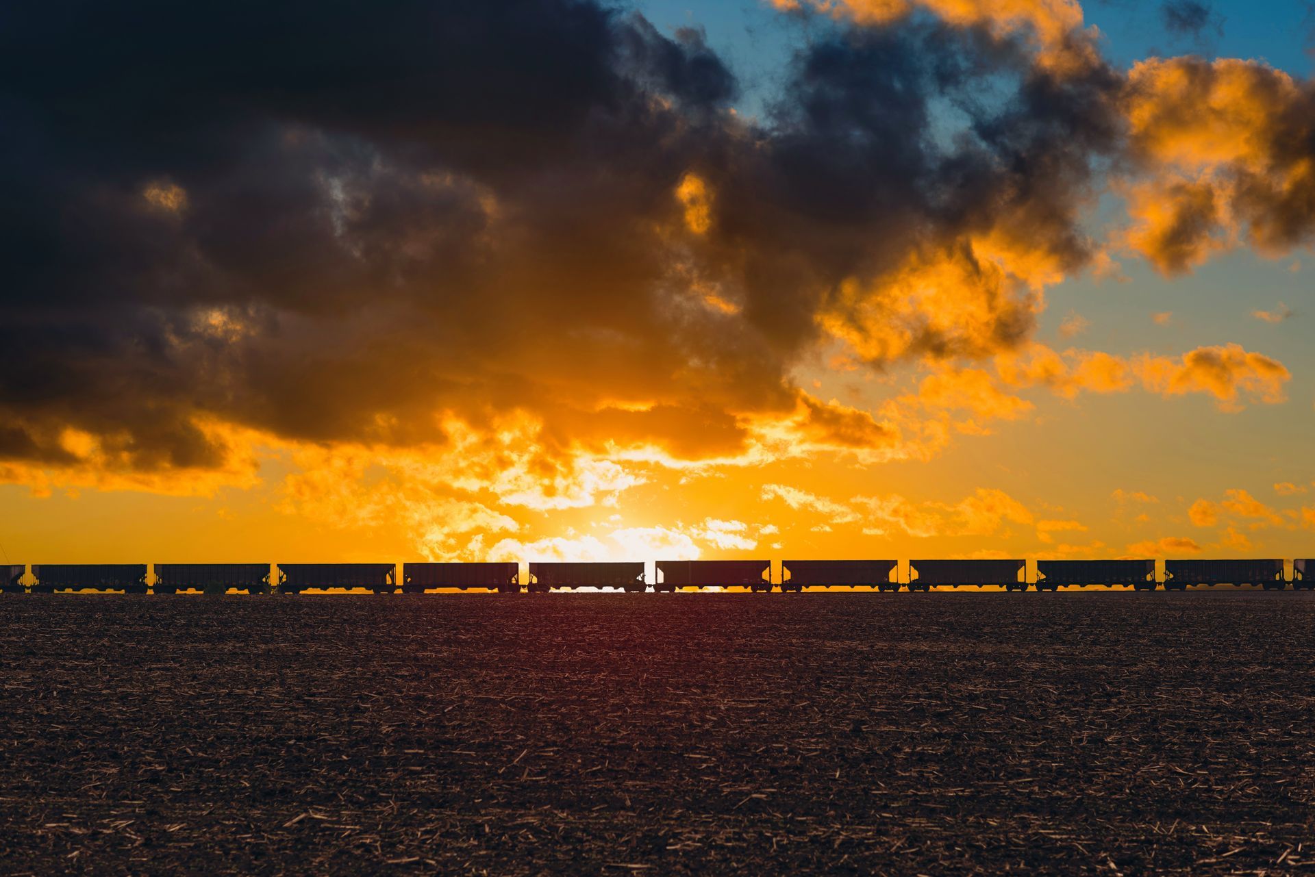 Train silhouetted against a sunset. Dark train cars on a horizon of farmland, under an orange and blue sky.