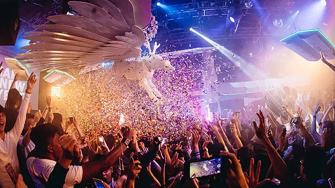 A crowd of people are dancing at a concert with confetti falling from the ceiling.