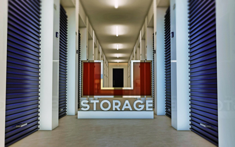 A long, brightly lit storage facility corridor with blue metal doors on each side and a red
