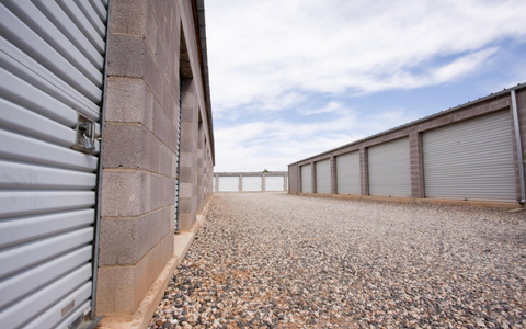 A gravel path between rows of grey storage units with roll-up doors under a blue sky.
