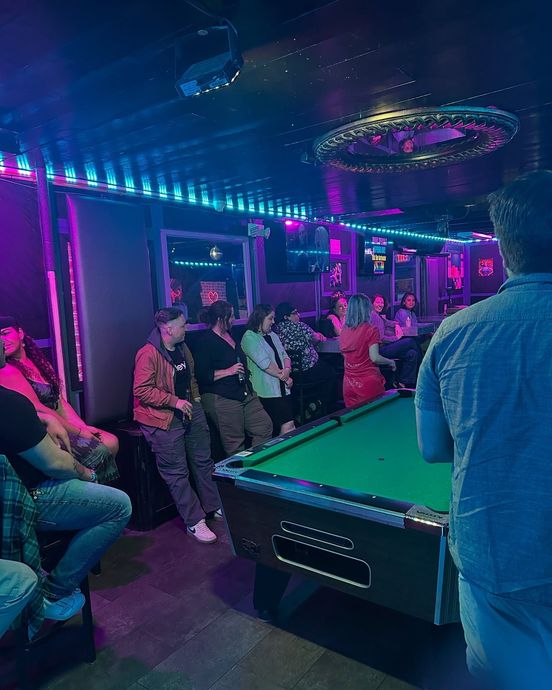 People in a bar with a pool table. Dim lighting with neon purple and blue hues.