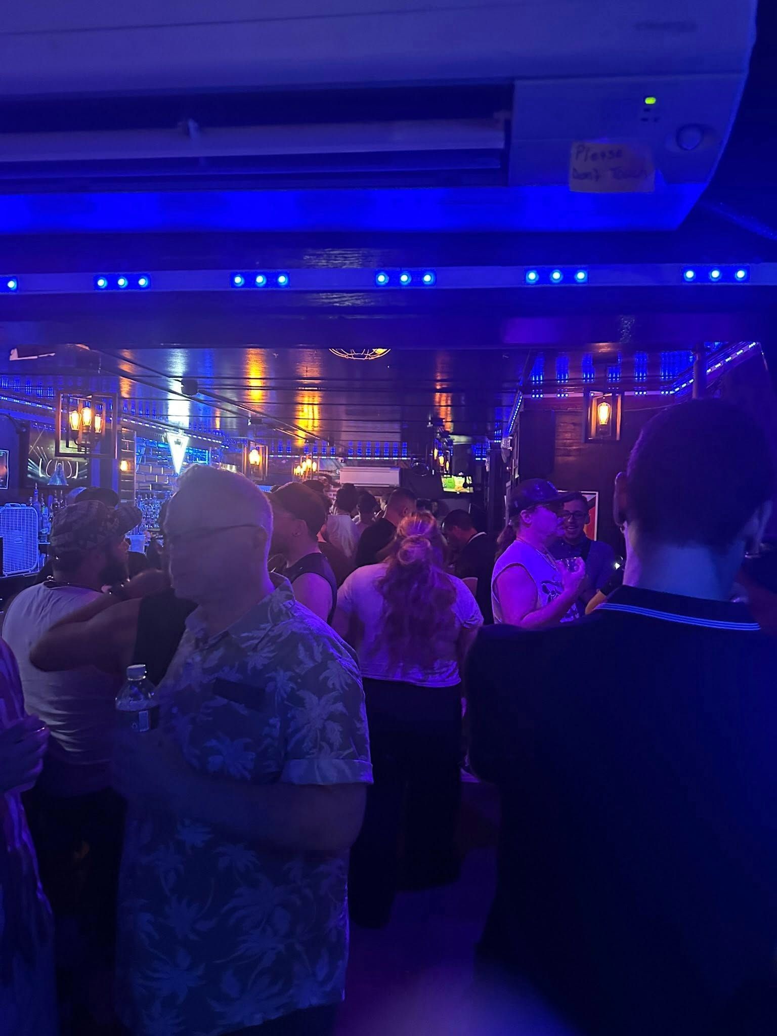 People in a dimly lit bar, some dancing. Blue and yellow lights illuminate the crowded space.