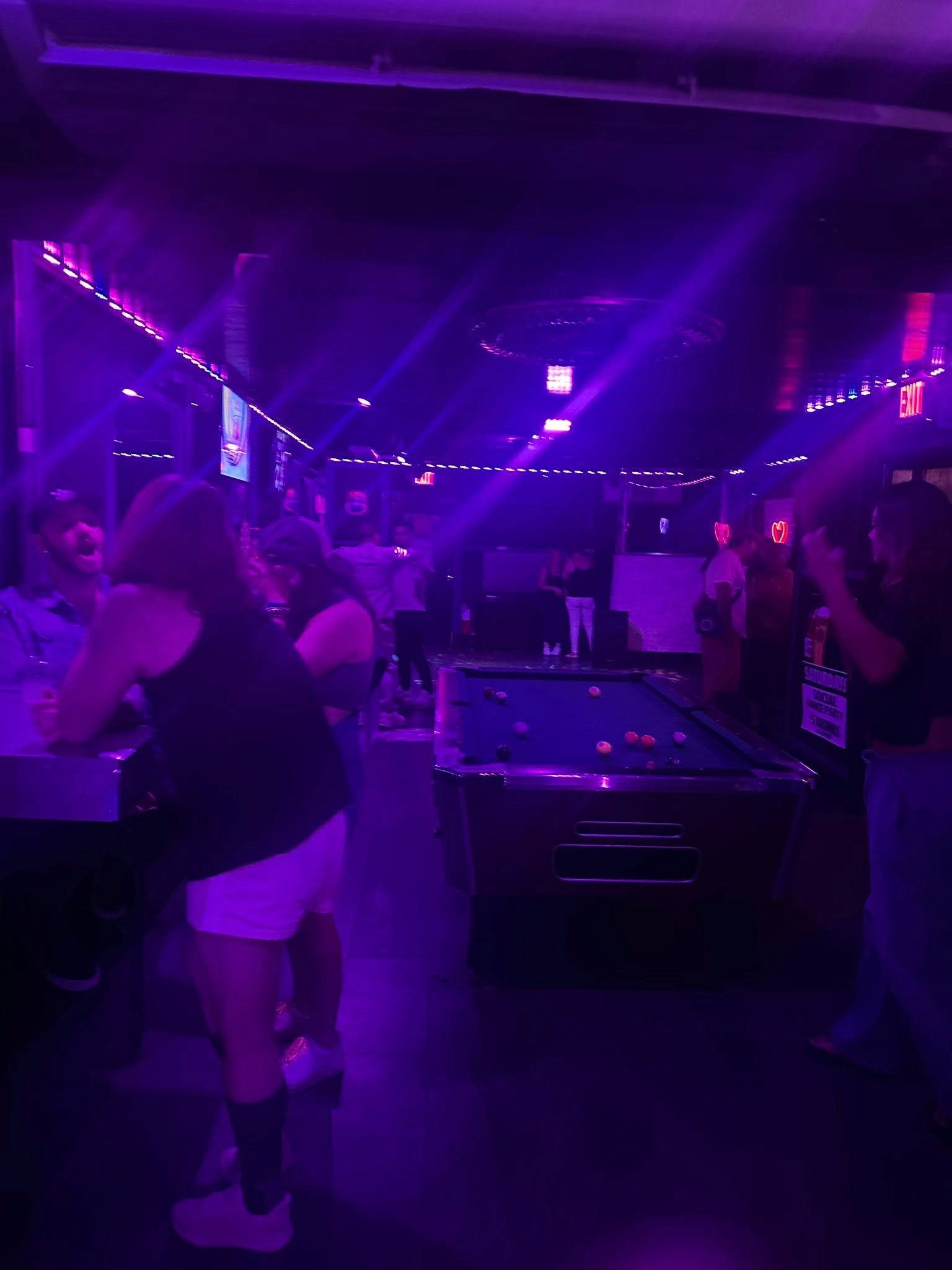 A dimly lit bar with purple lighting, pool table, and people socializing.
