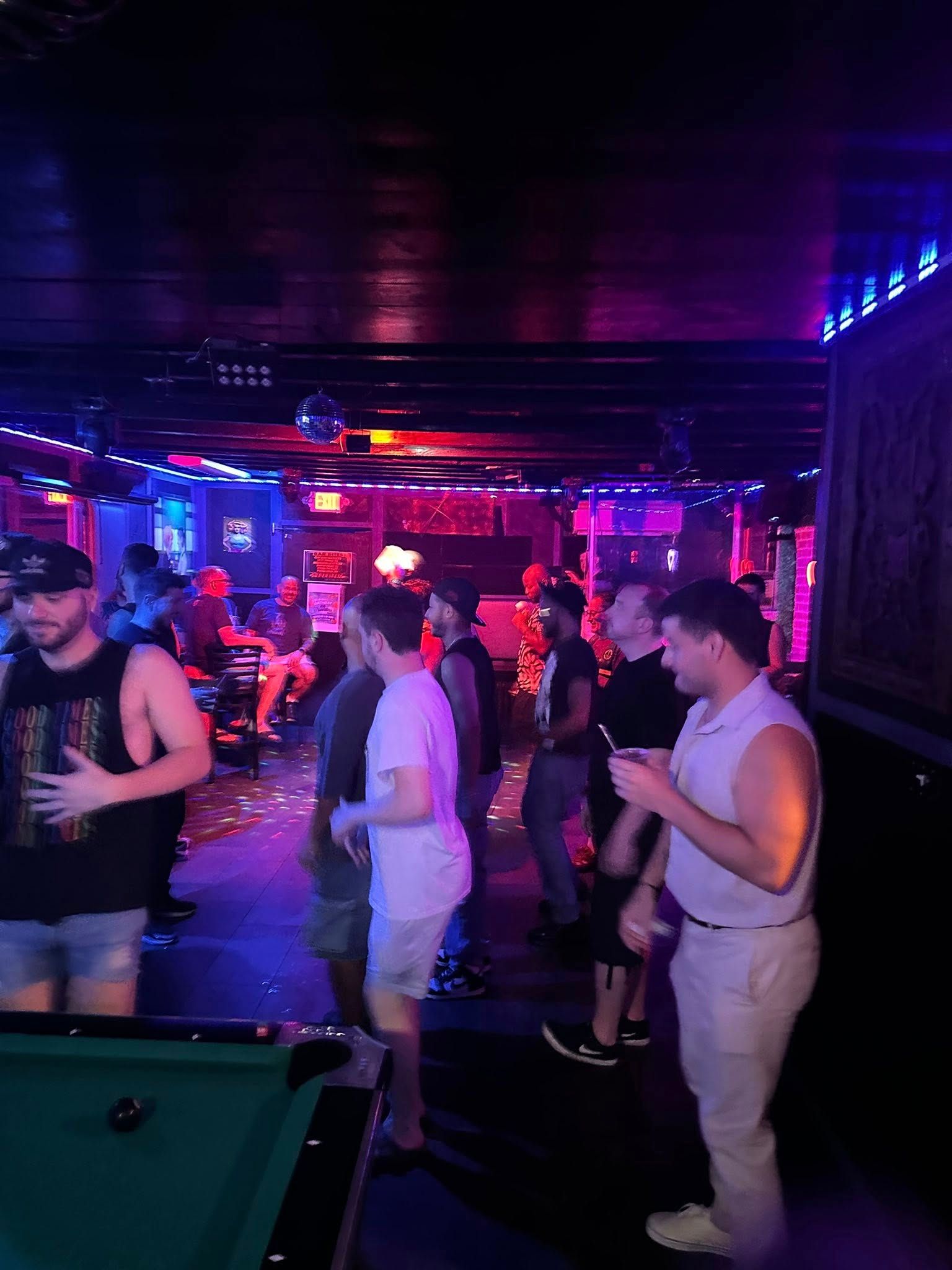 People in a dimly lit bar, some standing near a pool table. Purple and red lights illuminate the scene.