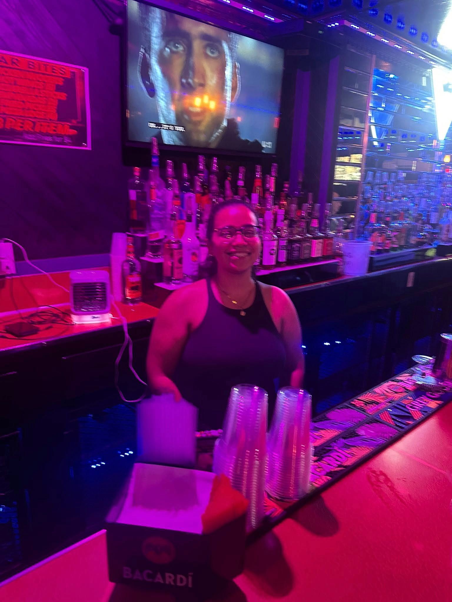 Woman bartender smiling behind a bar; bottles, glasses, and TV in the background. Purple and red lighting.