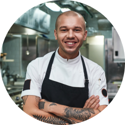 A man in an apron is smiling in a kitchen with his arms crossed