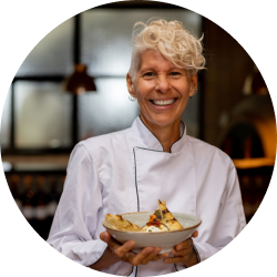 A woman in a chef 's uniform is smiling while holding a bowl of food