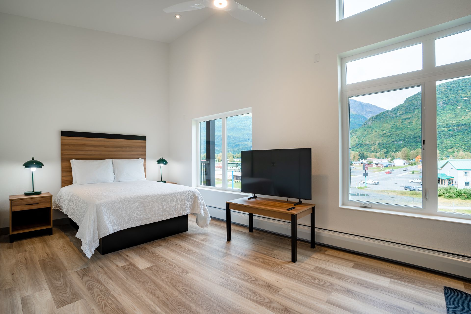 Bright hotel room with a bed, TV, and large windows overlooking mountains and a town.