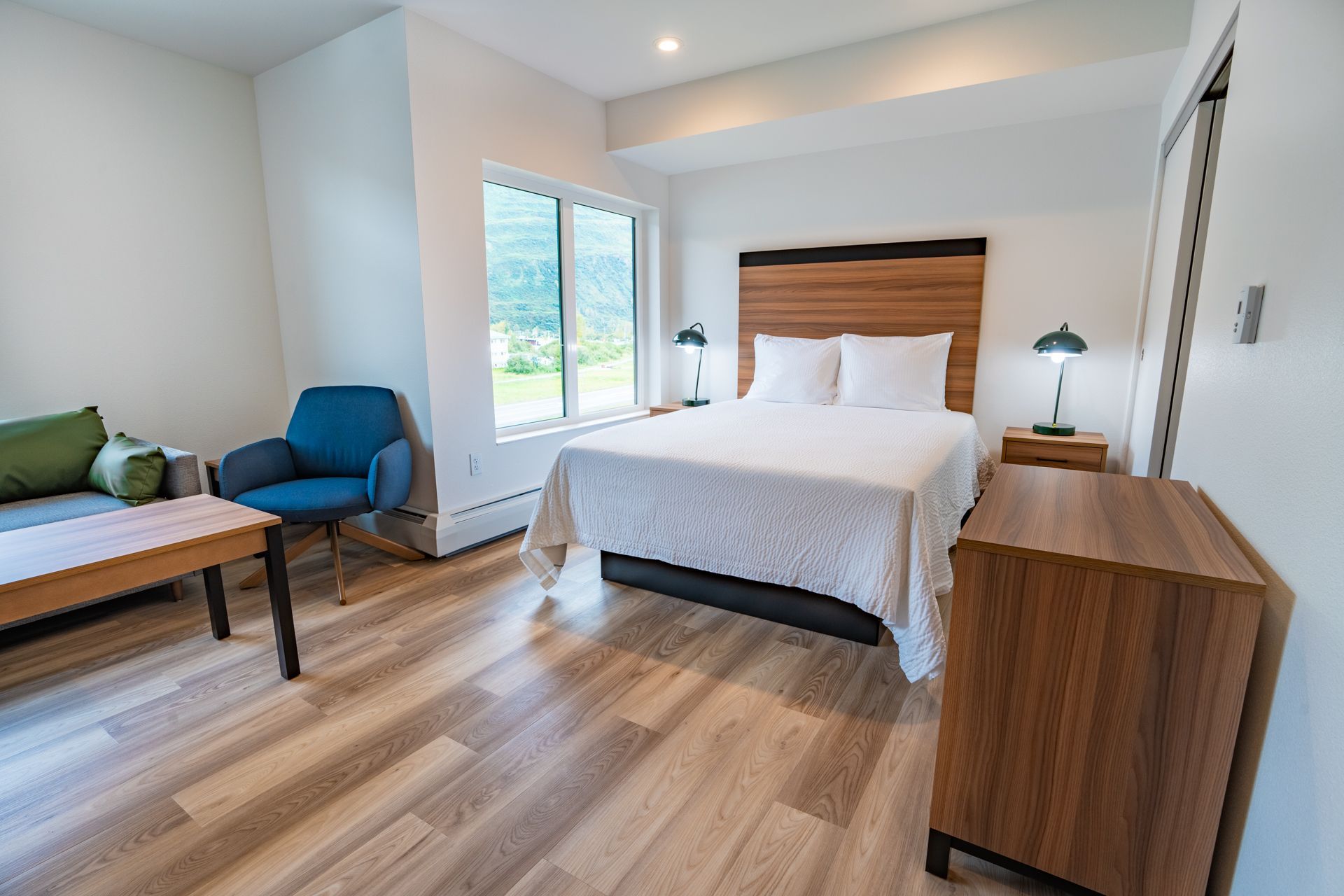 Bedroom with a bed, small sofa, blue chair, and a wood cabinet with a window overlooking a green landscape.