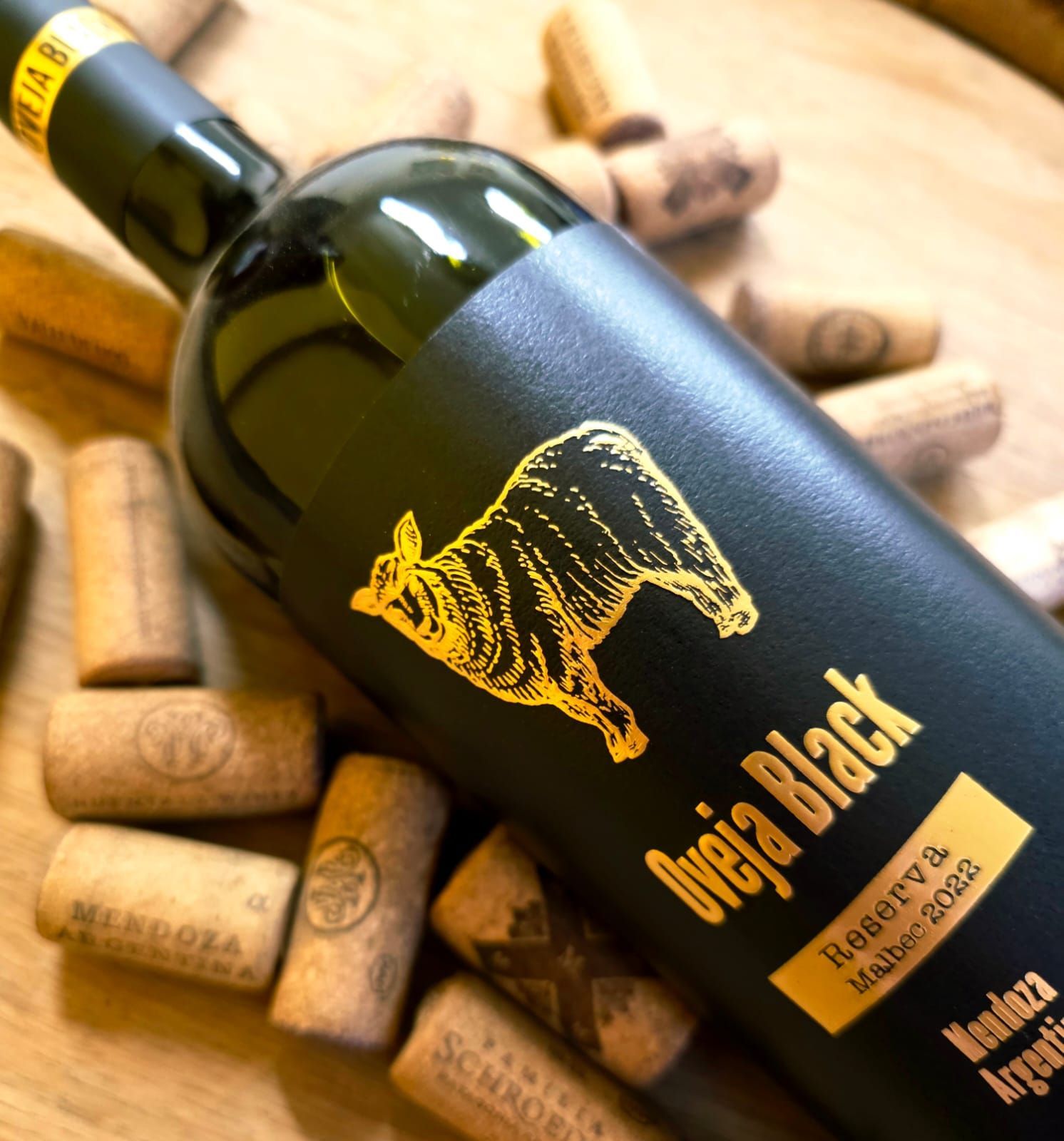 Una botella de vino Oveja Black Malbec rodeada de varios corchos de vino sobre una superficie de madera.