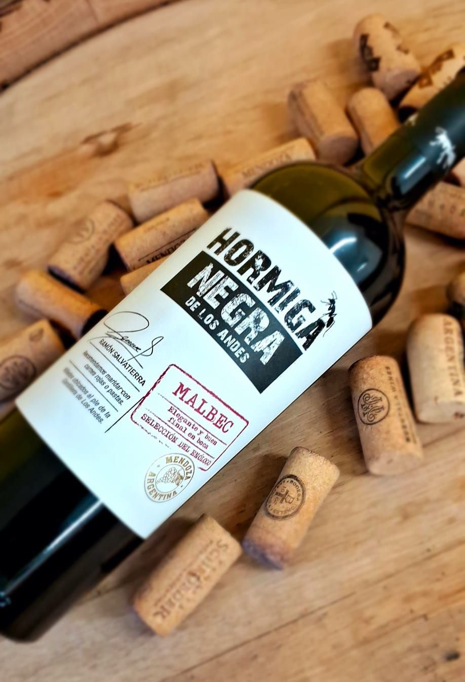 Una botella de vino Hormiga Negra Malbec rodeada de corchos de vino esparcidos sobre una superficie de madera.