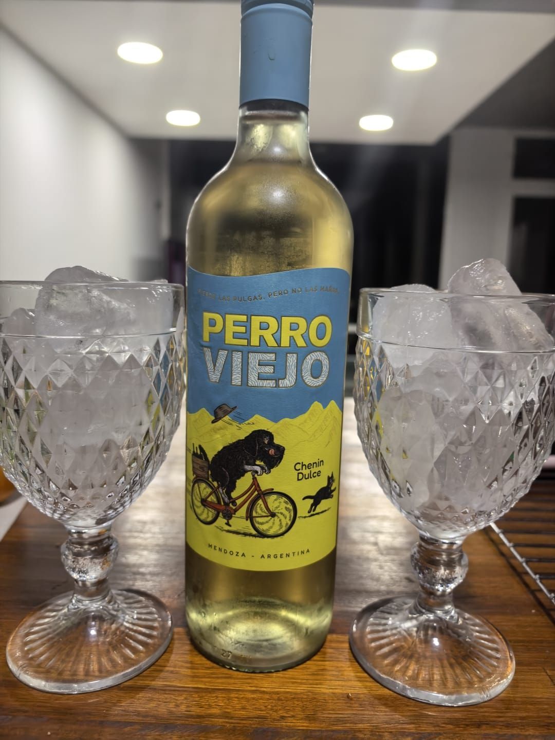 Una botella de vino blanco Perro Viejo reposa entre dos vasos llenos de hielo sobre una mesa de madera.