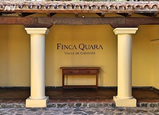 Letrero de Finca Quara entre dos columnas, con una mesita. Edificio marrón claro, suelo de piedra, Valle de Cafayate.