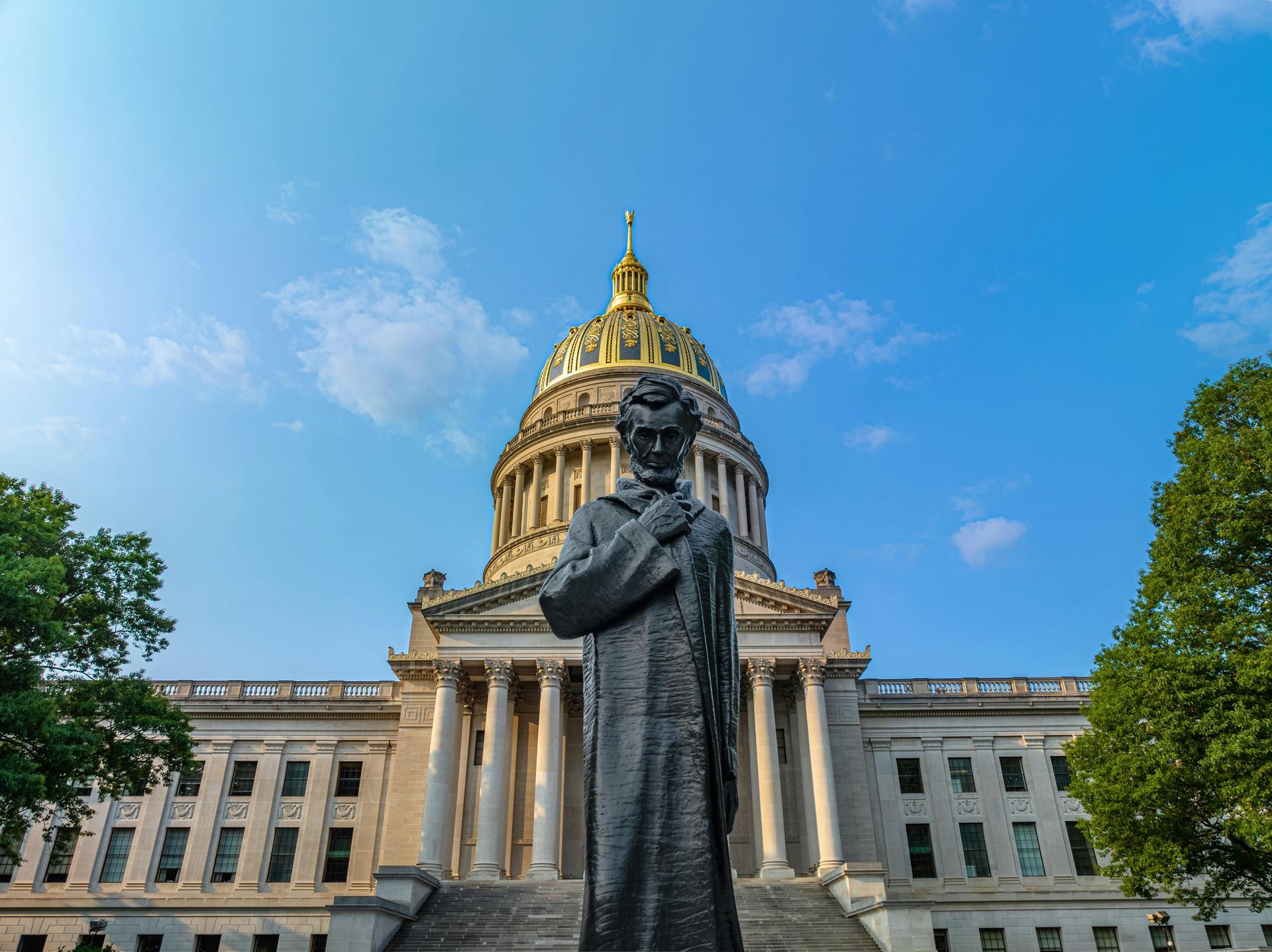 Statue of a man with arms crossed in front of West Virginia State Capitol building against a blue sky.