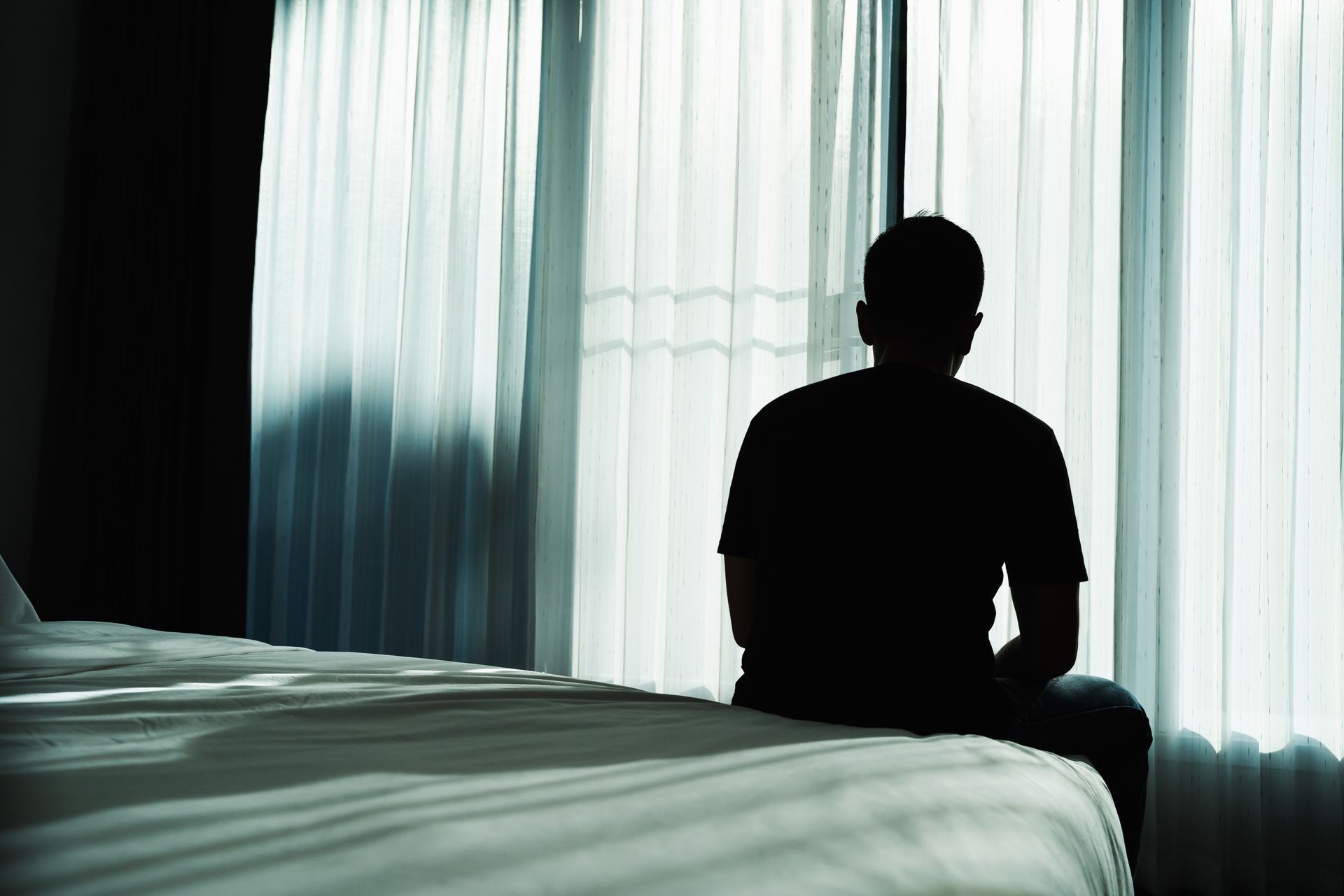 Silhouette of a person sitting on a bed, thinking, looking towards a window with sheer curtains.