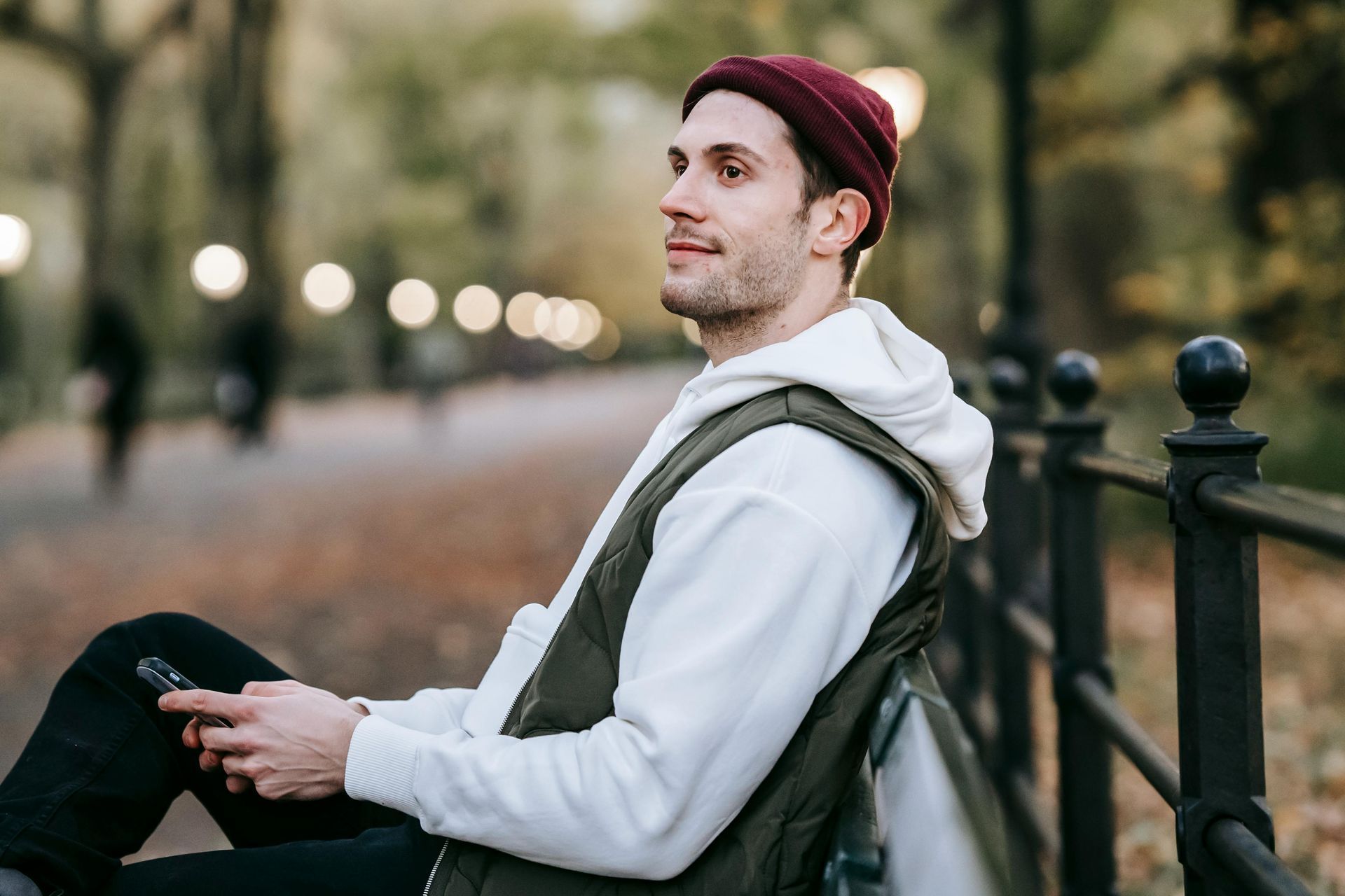 Man in a maroon beanie, white hoodie, and vest sits on a bench, looking to the side with a cell phone. Park setting.