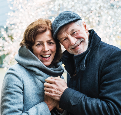 Smiling couple holding hands outdoors; woman in gray coat, man in blue coat and hat.