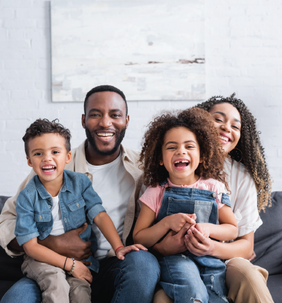 Family of four smiling, sitting on a couch.  Man, woman, and two children indoors.