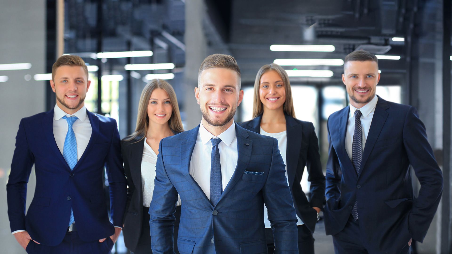 Five businesspeople in suits smiling in an office hallway.