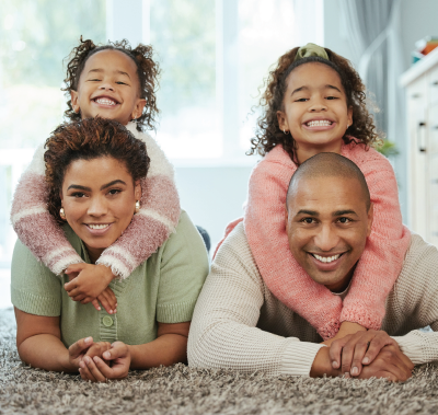 Family of four smiling, lying on carpet. Children are on parents' backs. Soft natural light.