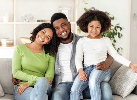 Family of three smiling on a couch; the parents embrace the child, who sits on father's lap.