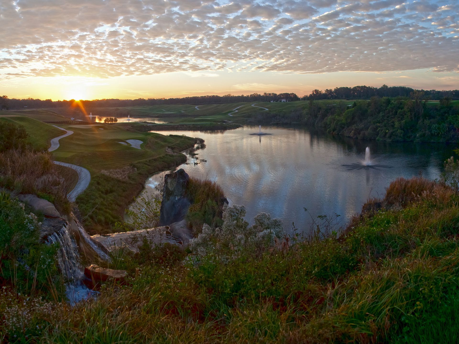 A lake with a fountain in the middle of it at sunset.