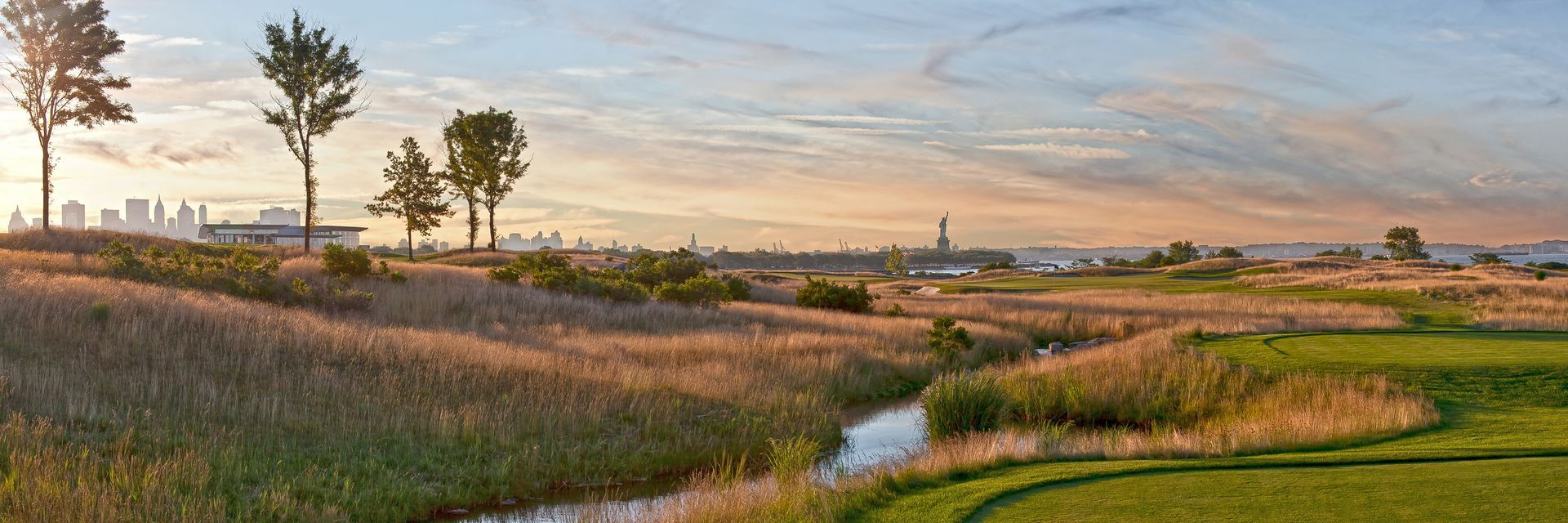 A lush green field with trees and a river in the foreground and a city in the background.