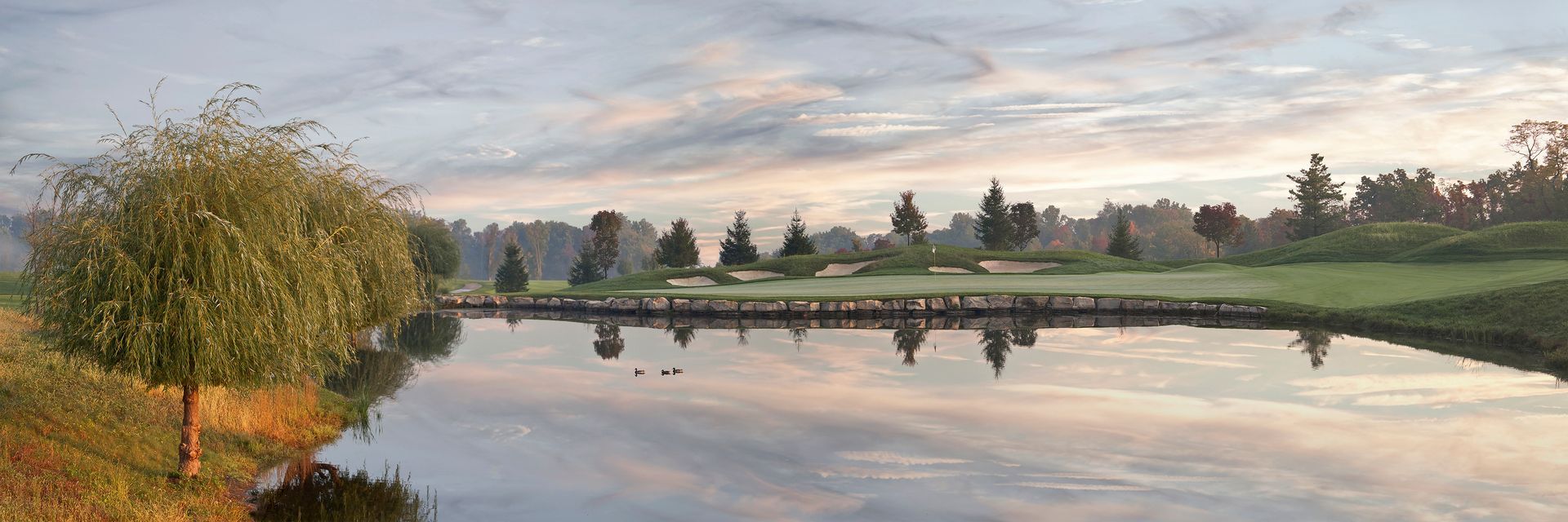 A painting of a river with trees on the shore and a golf course in the background.