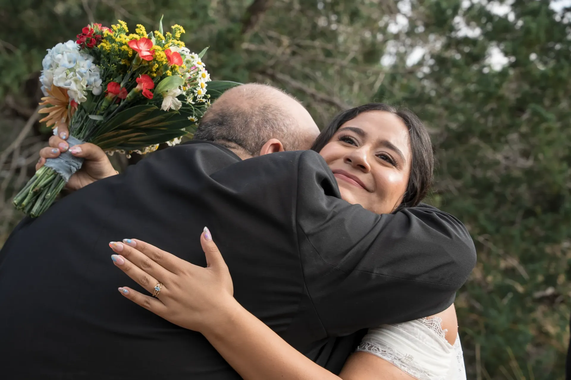 A person in a wedding dress hugs another person in a dark suit, holding a colorful bouquet in their left hand.