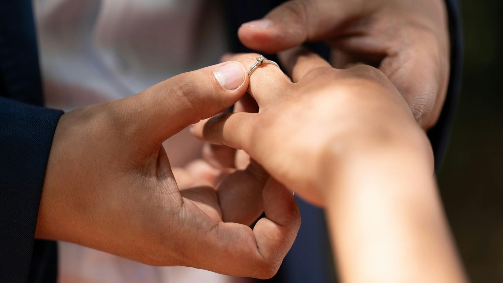 A person in a dark suit jacket gently slides a delicate ring onto another person's finger during a wedding ceremony.