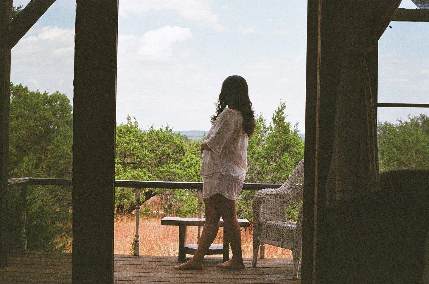 A person in light-colored clothing stands on a rustic porch, looking out over a view of lush green trees and a field.