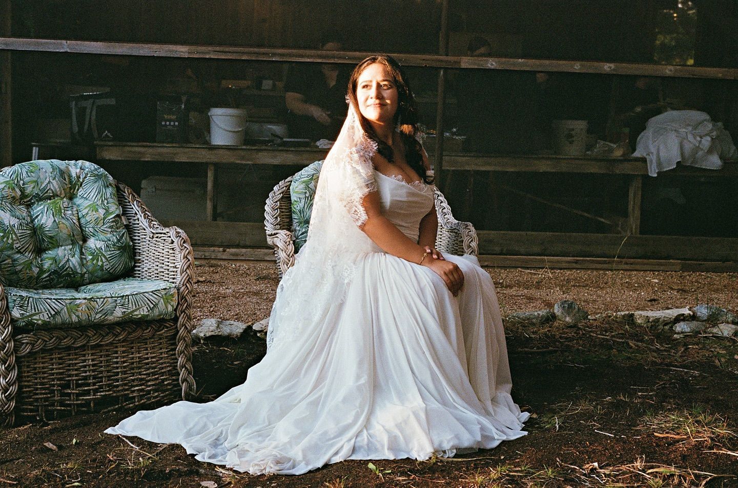 A person in a white wedding gown and veil sits on a wicker chair outdoors in front of a rustic wooden structure.