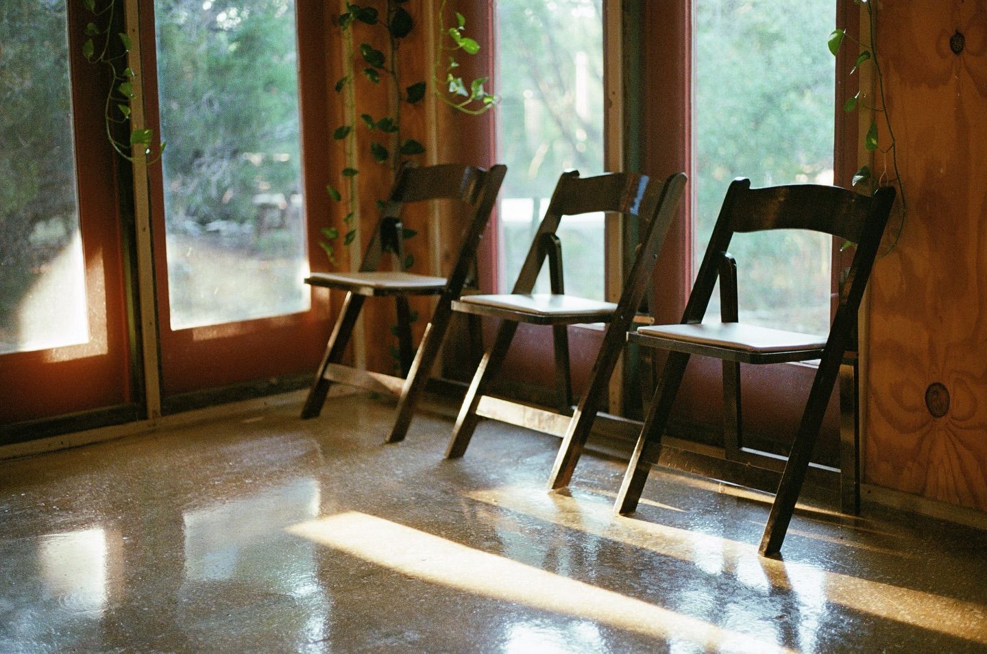 Three dark wooden folding chairs sit in a row on a reflective, speckled floor in front of large glass windows.