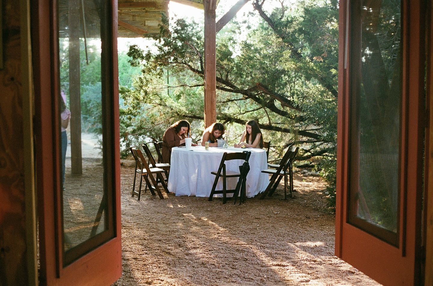 Three people sit at a round, white-clothed table under trees, viewed through an open doorway.
