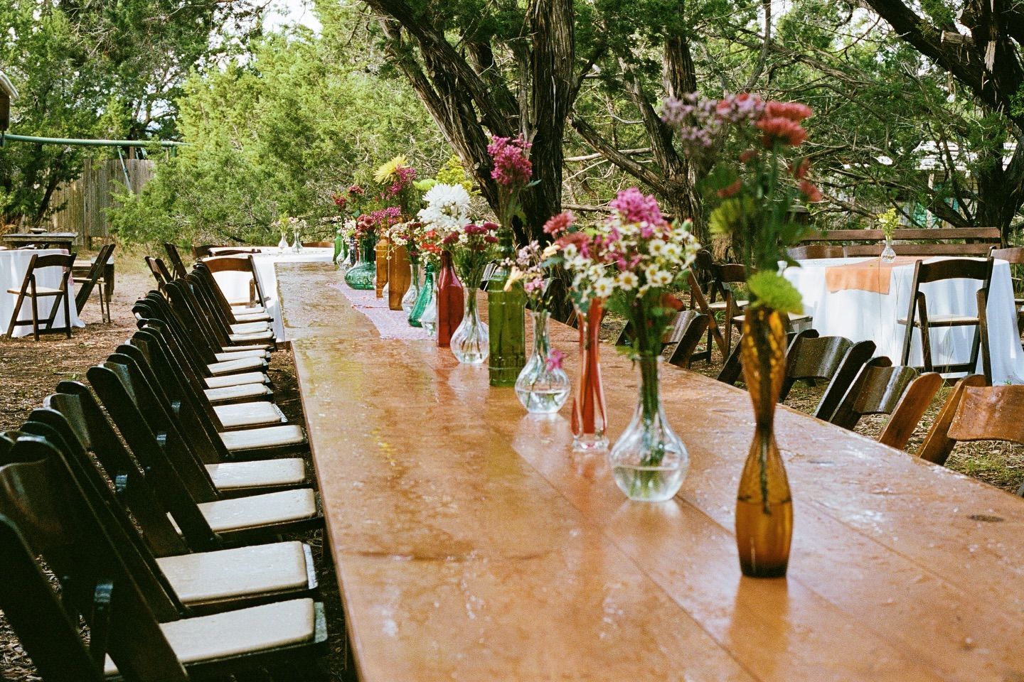 A long wooden banquet table outdoors, set for an event with colorful flower vases and rows of folding chairs.