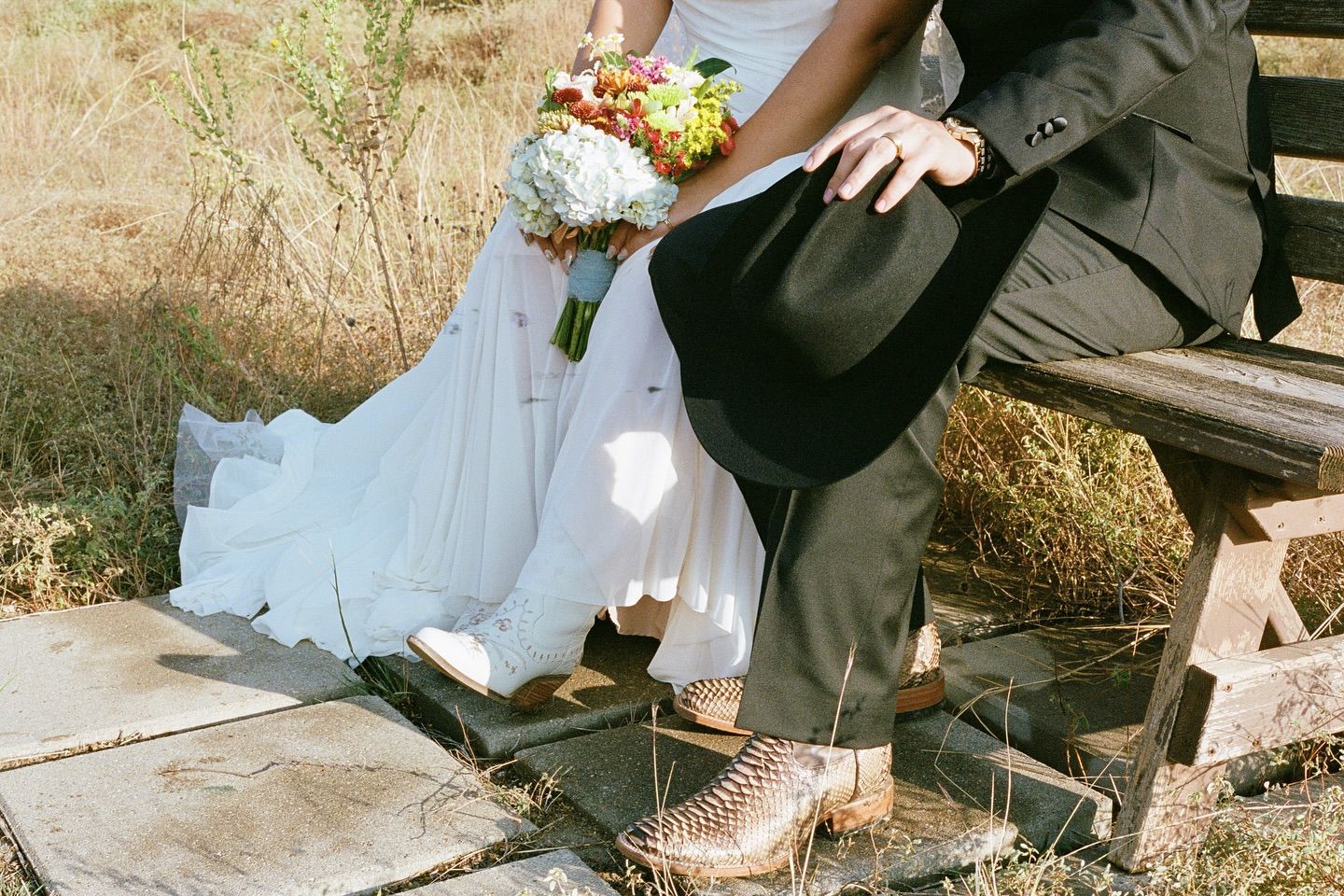 A bride and groom wearing cowboy boots sit on a wooden bench outdoors, holding a bouquet of colorful flowers.