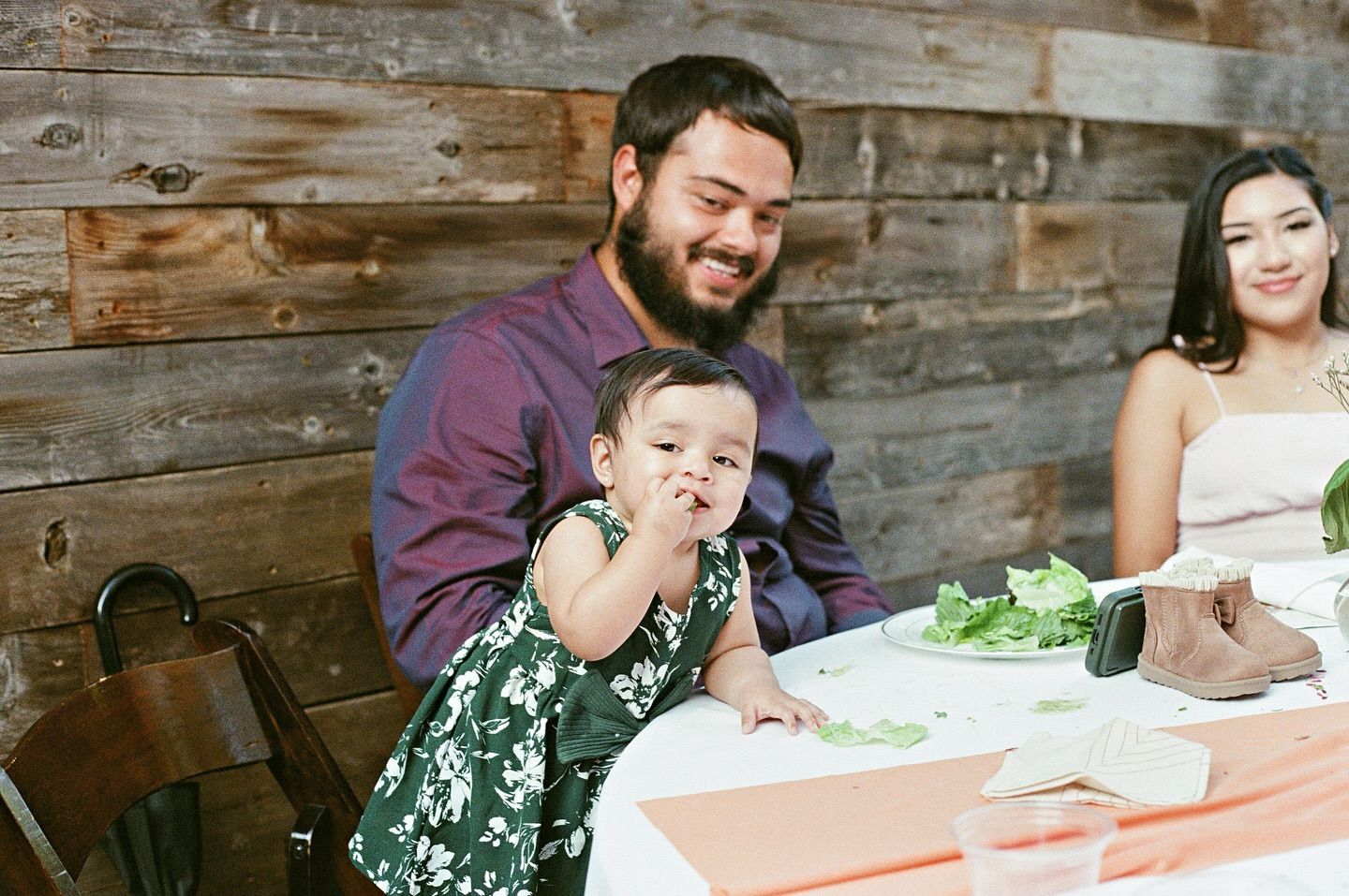 A person holding a baby in a green floral dress sits at a table near a woman against a wood-paneled wall.