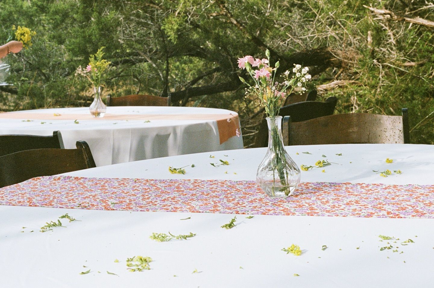 Two round tables with white tablecloths and patterned runners, set outdoors with clear vases holding floral arrangements.