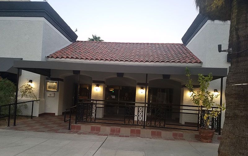 Exterior of a building with a red-tiled roof, arched doorways, and a black awning. A ramp and railings provide accessibility.
