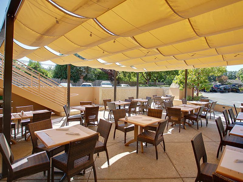 Outdoor restaurant seating under a tan awning. Tables and chairs are set for diners; parking area in background.