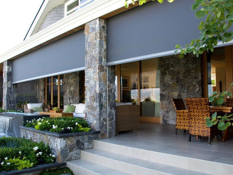 Gray outdoor roller shades on a stone house patio, covering windows. Steps lead down to a dining area with wicker furniture.
