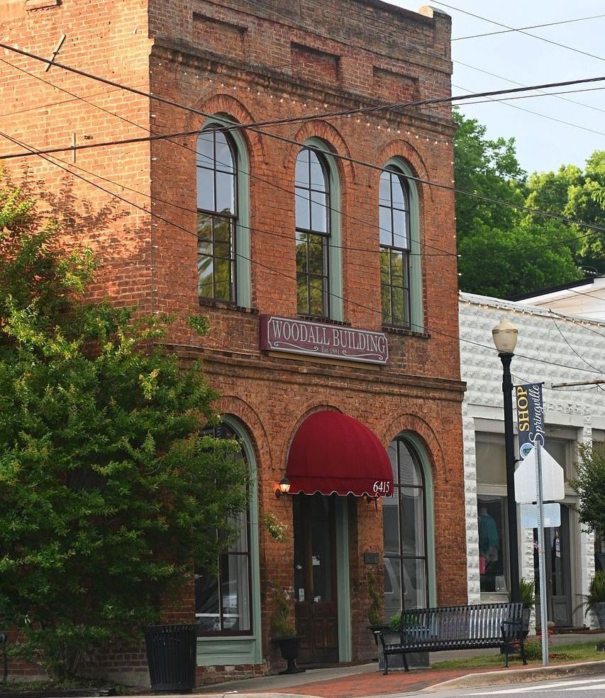 A three-story brick building with arched windows and a red awning, fronted by a street and bench.