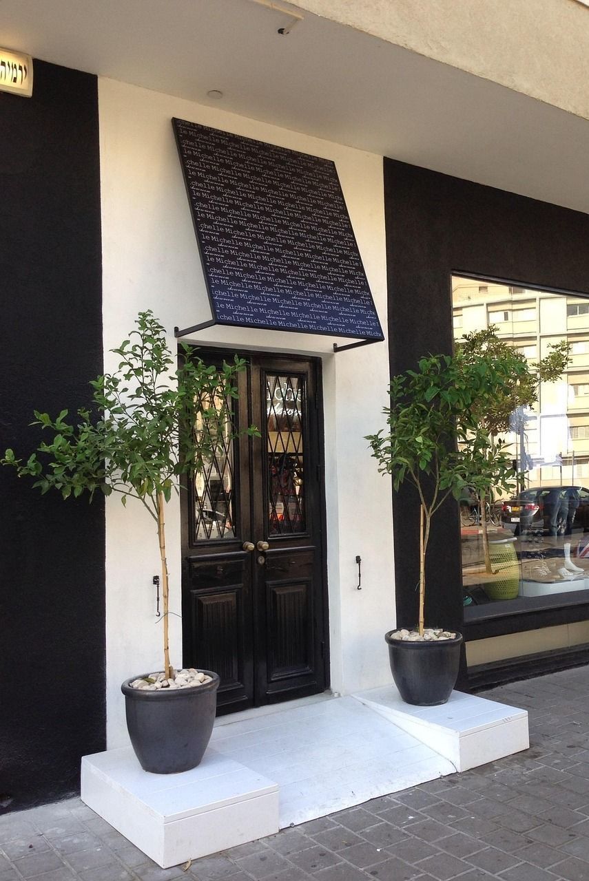 Black double doors with textured awning, flanked by potted trees on a white platform, set against a black and white building facade.