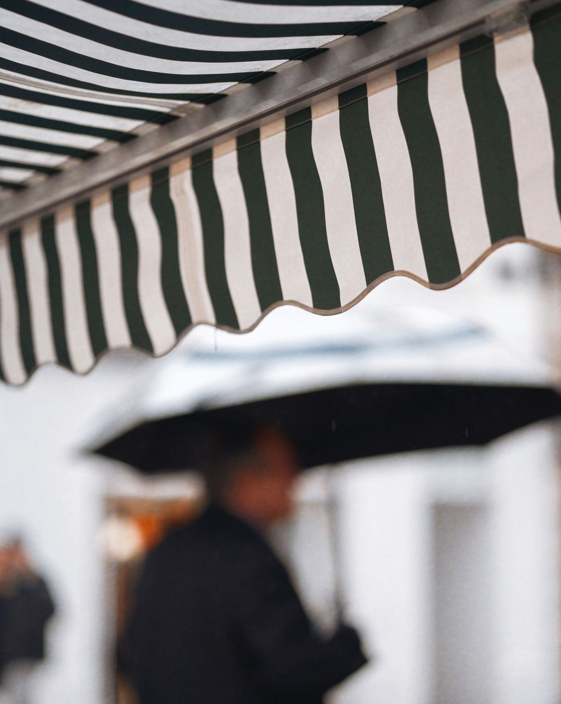 Green and white striped awning over a person with an umbrella, blurred background of buildings.